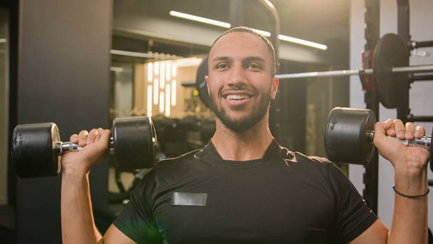 Happy smiling African American sportsman doing power exercise at gym with sport equipment bodybuilder man lifting heavy weights dumbbells workout male fitness athlete training shoulders hands muscle - Foto, Bild