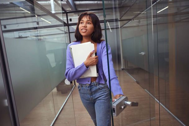 Joyful woman employee with braces opening door while holding work items in modern room - Photo, image