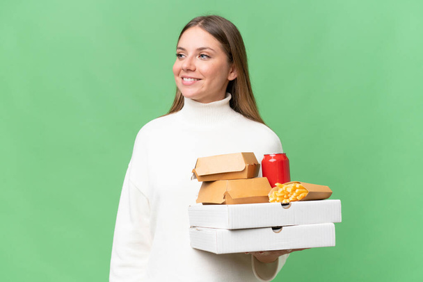 Νέα όμορφη ξανθιά γυναίκα κρατώντας takeaway τροφίμων πάνω από απομονωμένη φόντο κοιτάζοντας στο πλάι και χαμογελώντας - Φωτογραφία, εικόνα