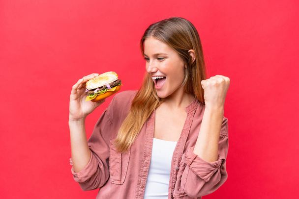 Joven mujer caucásica sosteniendo una hamburguesa aislada sobre fondo rojo celebrando una victoria - Foto, imagen