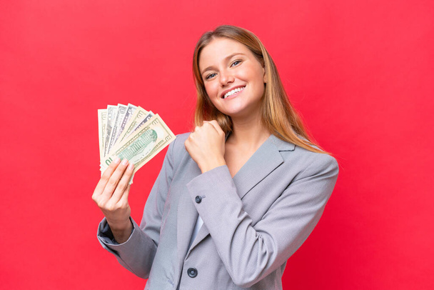 Joven mujer caucásica de negocios sosteniendo el dinero aislado sobre fondo rojo celebrando una victoria - Foto, Imagen