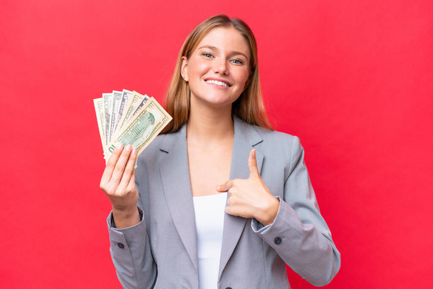 Giovane donna caucasica d'affari che tiene i soldi isolati su sfondo rosso con espressione facciale a sorpresa - Foto, immagini