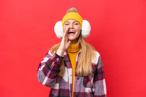 Young beautiful woman wearing winter muffs isolated on red background shouting with mouth wide open - Foto, imagen