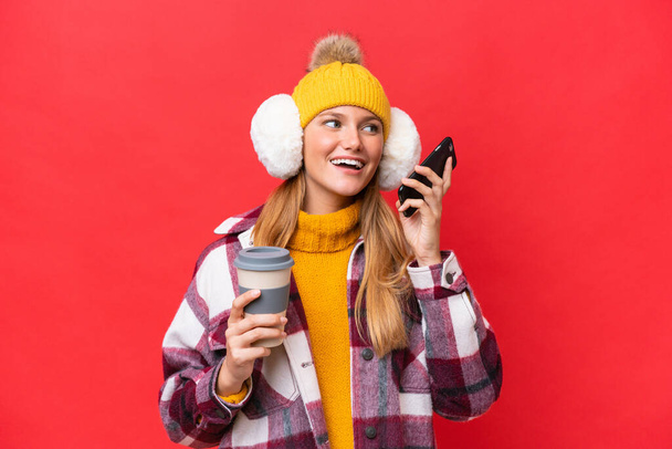 Mujer hermosa joven con manguitos de invierno aislados sobre fondo rojo sosteniendo café para llevar y un móvil - Foto, imagen