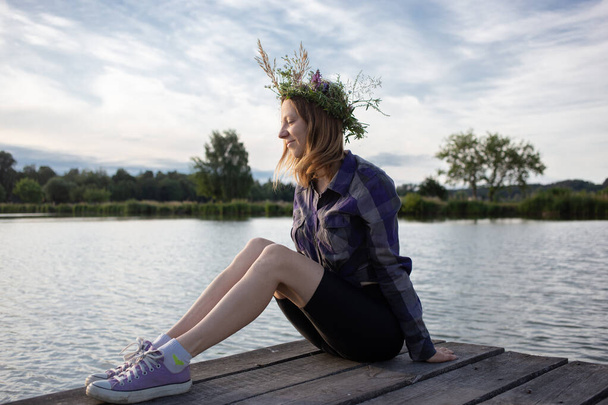 lifestyle portrait of cute woman in wreath of wildflowers. sitting on wooden pier by water. romantic mood. Holiday for nature lovers. Traditions of the holiday of Ivan Kupala. Enjoy the summer, relax - Photo, Image