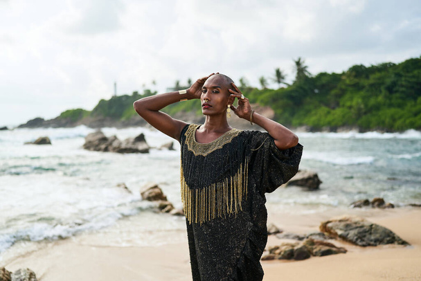 Epatage lgbtq black male posing with hands up on camera on scenic ocean beach. Non-binary ethnic fashion model in long posh dress wears jewellery stands gracefully on sea shore and a lighthouse. - Photo, Image