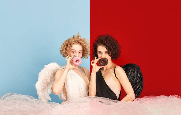 Portrait of beautiful women in costumes of angel and demon posing with donuts against blue red background. Strawberry or chocolate. Concept of emotions, comparison of being, creativity, beauty - Foto, imagen