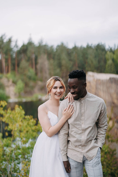 Happy interracial couple newlyweds stands and laughs cheerfully against background of lake and forest. Concept of love relationships and unity between different human races. - 写真・画像