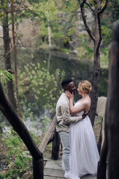 Happy interracial couple newlyweds stands on wooden bridge, embraces and smiles against background of lake and forest. Concept of love relationships and unity between different human races. - Photo, Image