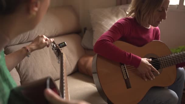 dos mujeres de unos cuarenta años practicando la guitarra en el sofá, afinando sus guitarras con un afinador y un teléfono móvil. Guitarra clásica y acústica - Metraje, vídeo