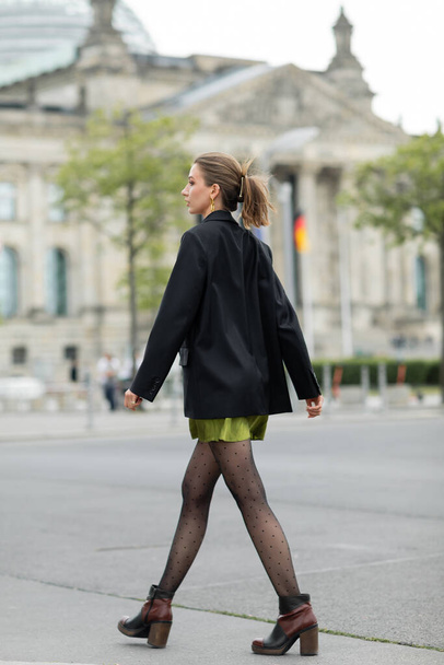 Πλευρική άποψη της κομψής νεαρής γυναίκας με μαύρο σακάκι και μπότες κοιτάζοντας μακριά, ενώ το περπάτημα στο Βερολίνο - Φωτογραφία, εικόνα