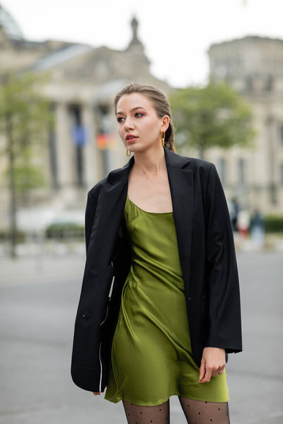 Fashionable and pretty young fair haired woman in green sink dress and black jacket in Berlin - Photo, Image