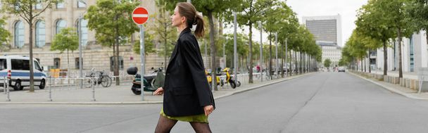 Вид сбоку на элегантную и стильную молодую женщину в платье и черной куртке, идущую по Берлину, баннер  - Фото, изображение