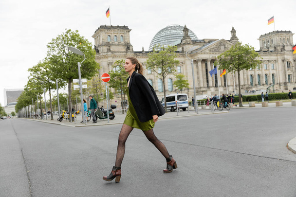 Pełna długość młodej i jasnowłosej kobiety w jedwabnej sukience, kurtce i butach spacerujących po drodze w Berlinie - Zdjęcie, obraz