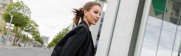 Pozitív fiatal szőke nő fekete blézerben, félrenéz, miközben Berlinben sétál, banner  - Fotó, kép