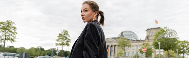 Joven mujer de pelo justo de moda en chaqueta negra mirando hacia otro lado mientras está de pie en Berlín, pancarta  - Foto, Imagen