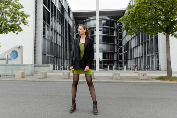 Volle Länge der trendigen jungen Frau in Stiefeln und schickem Blazer, die auf der Straße in Berlin wegschaut - Foto, Bild