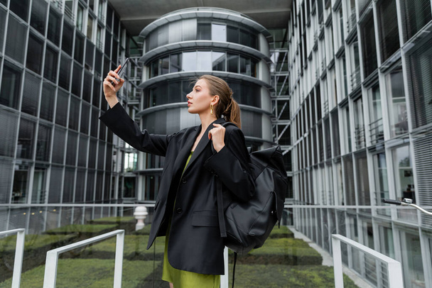 Seitenansicht einer modischen jungen Frau in Jacke, die einen Rucksack hält und ein Selfie mit dem Smartphone macht   - Foto, Bild