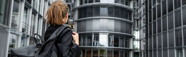 Visão traseira da jovem mulher de cabelos bonitos em jaqueta segurando mochila e tirando foto, bandeira  - Foto, Imagem