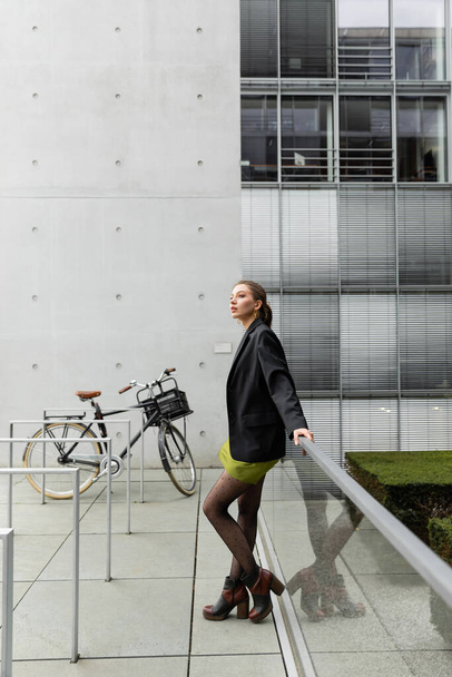 Stylische und hübsche junge Frau in schwarzer Jacke und Seidenkleid steht neben Fahrrad in Berlin  - Foto, Bild