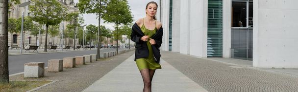 Módní žena v hedvábných šatech a černém blejzru při pohledu do kamery v Berlíně, Německo, prapor  - Fotografie, Obrázek