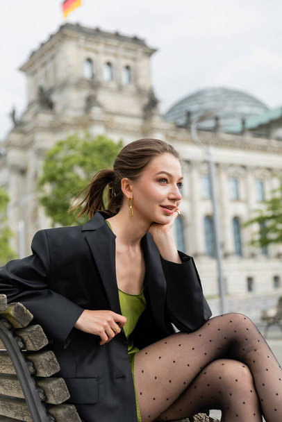 Verträumte junge Frau in Jacke, Kleid und Strumpfhose schaut weg, während sie auf einer Bank in Berlin sitzt - Foto, Bild