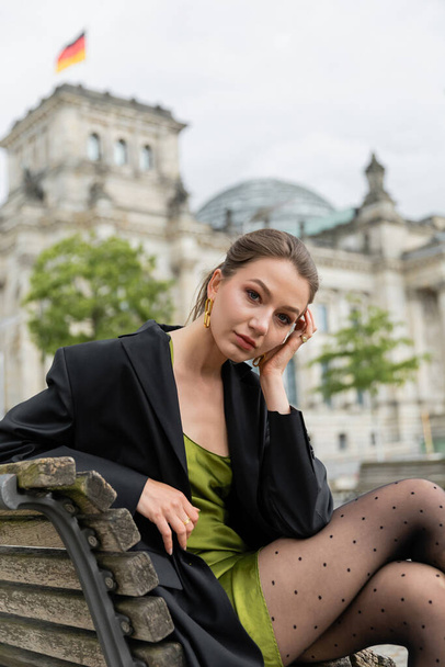 Portret van een stijlvolle vrouw in jasje en zijden jurk kijkend naar de camera en zittend op een bankje in Berlijn - Foto, afbeelding