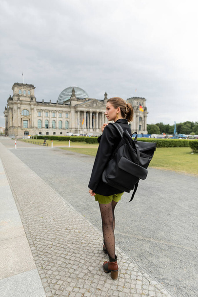 стильна жінка в куртці і сукні тримає рюкзак під час прогулянки біля будівлі Рейхстагу в Берліні - Фото, зображення