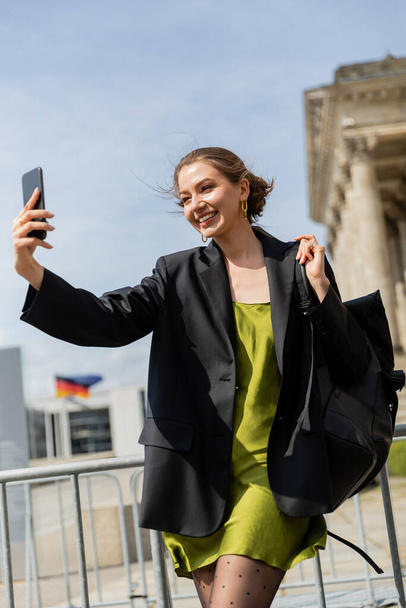 Donna allegra in giacca e vestito di seta che tiene lo zaino e si fa selfie a Berlino, Germania  - Foto, immagini