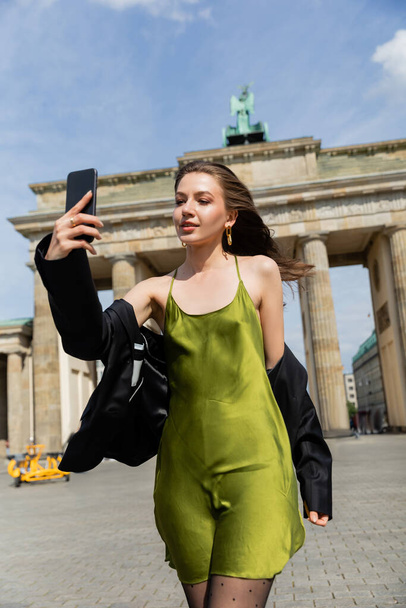 Модная женщина в пиджаке и шелковом платье делает селфи возле Бранденбургских ворот в Берлине - Фото, изображение