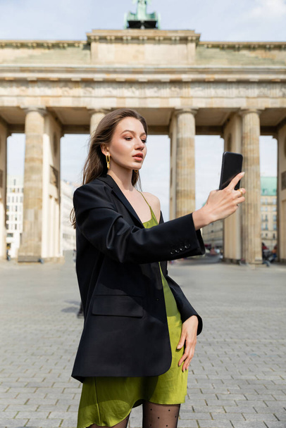ベルリンのブランデンブルク門の近くでセルフィーを取るジャケットと絹のドレスでエレガントなファッショナブルな女性 - 写真・画像