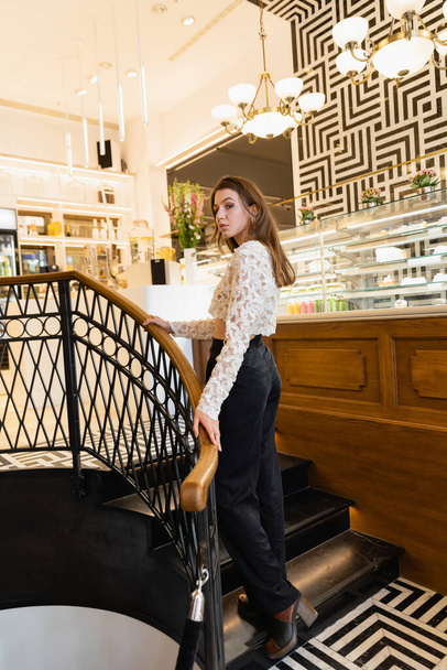 Young and stylish fair haired woman in lace top and pants standing in modern cafe in Berlin - Photo, Image