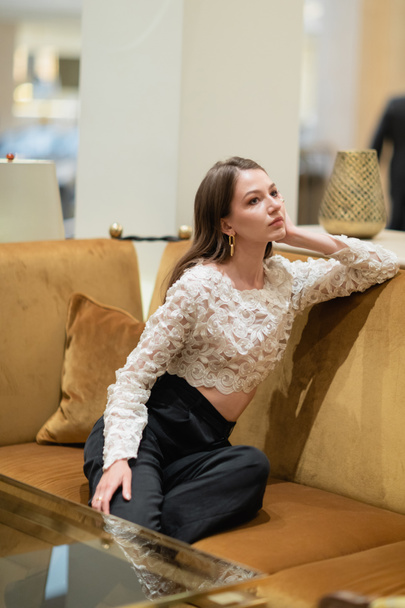 relaxed fair haired woman in lace top and pants sitting on couch in hotel lobby of Berlin - Photo, Image