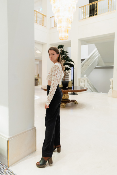 Полная длина модной женщины в кружевном топе, брюках и сапогах, смотрящей на камеру в берлинском отеле - Фото, изображение