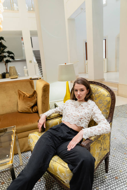 young and stylish woman in lace top and pants looking at camera while sitting on armchair  - Photo, Image