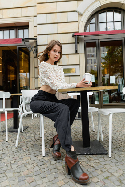 стильная женщина в кружевном топе и брюках, держащих кофе и сидящих за столом открытого кафе в Берлине  - Фото, изображение