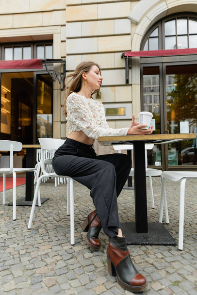 мечтательная женщина в кружевном топе и брюках, держащих кофе и сидящих за столом открытого кафе в Берлине  - Фото, изображение