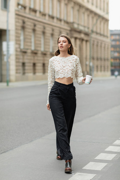 mujer con estilo en la parte superior de encaje y pantalones de cintura alta sosteniendo café para ir a la calle en Berlín, Alemania  - Foto, Imagen