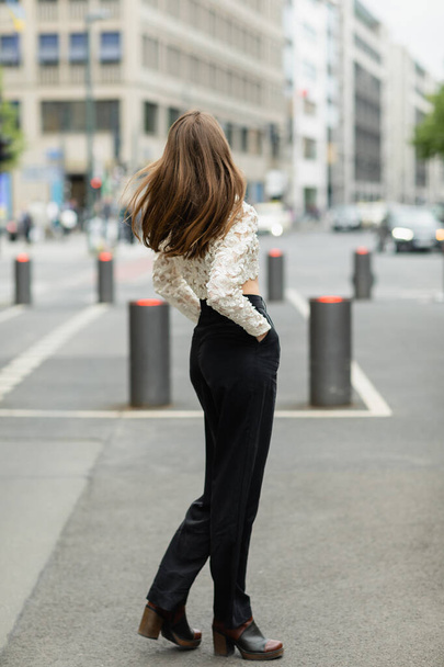 Back view of young fair haired woman in lace top and high waist pants standing on street in Berlin - Photo, Image