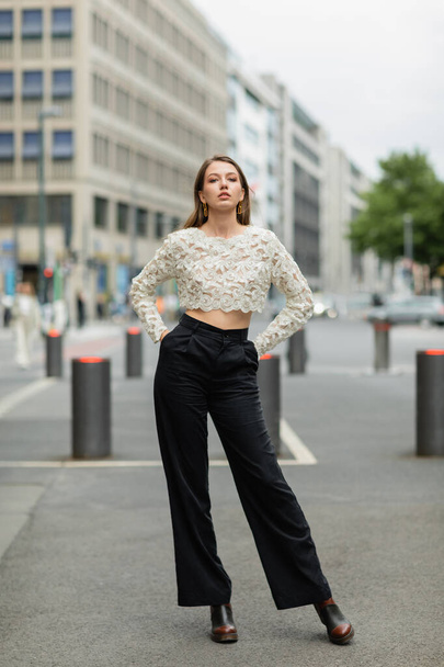 Młoda kobieta pozowała w koronkowej bluzce i wysokich spodniach w pasie stojąc na ulicy w Berlinie, Niemcy - Zdjęcie, obraz
