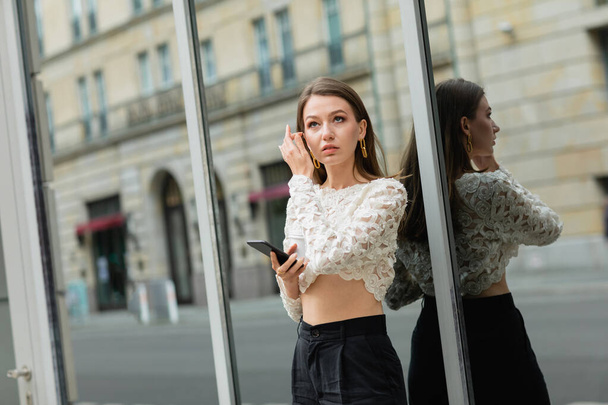 pensive young woman holding smartphone while standing near building on street in Berlin, Germany  - Photo, Image