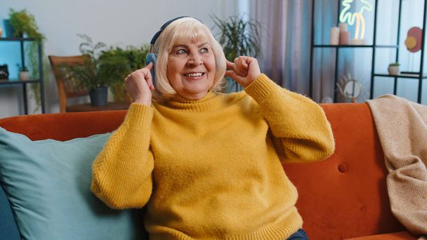 Счастливая старушка в беспроводных наушниках сидит на диване дома, выбирая любимую энергетическую диско-музыку. Отдых в выходные дни - Фото, изображение