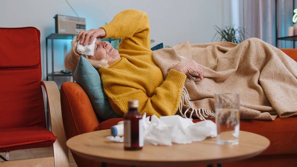 Grandmother ill suffering from cold or allergy pain lying on couch in living room at home. Sick mature woman with towel on head blows sneezes wipes snot into napkin. Coronavirus quarantine pandemic - 写真・画像