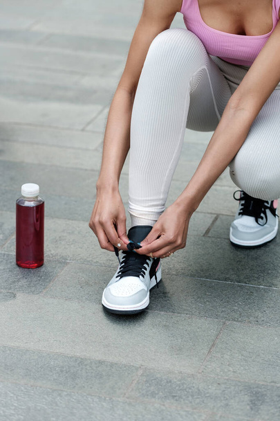 Unrecognizable fitness woman tying her shoelaces outdoors on grey rough floor tiles. She is wearing leggings and a pink sport bra. There is a bottle of energy drink on the floor. - Foto, Imagem