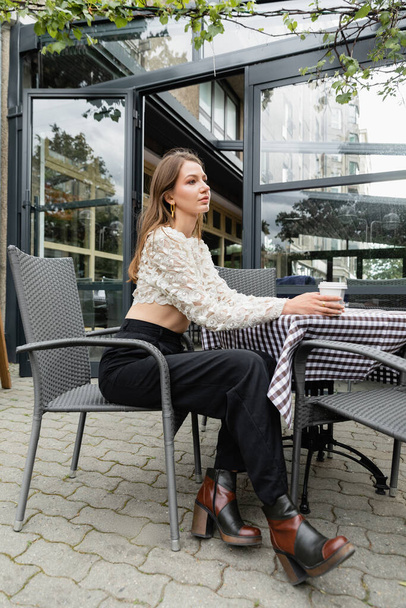 Модная молодая женщина держит кофе, чтобы пойти, сидя за столом на террасе возле кафе в Берлине - Фото, изображение