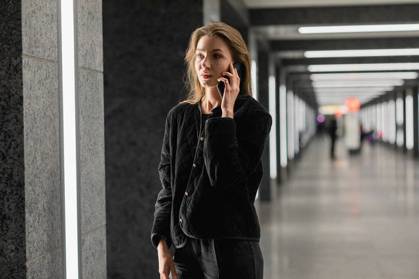 Stílusos nő fekete kabátban beszél okostelefonon, miközben bent áll a modern épületben - Fotó, kép