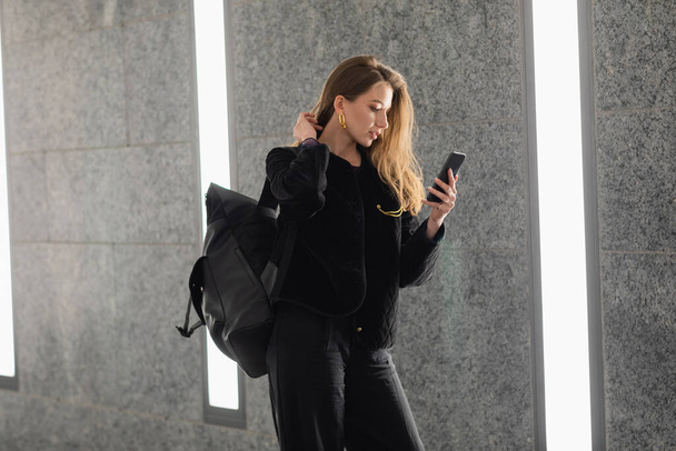 Stylová žena v bundě s batohem dotýkající se vlasů a pomocí smartphonu v blízkosti zářivek  - Fotografie, Obrázek