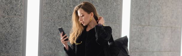 Donna alla moda in giacca con zaino utilizzando il cellulare mentre in piedi vicino al muro di cemento grigio, banner - Foto, immagini