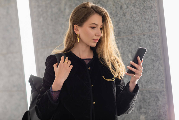 νεαρή γυναίκα με μαύρο σακάκι κρατώντας σακίδιο και χρησιμοποιώντας smartphone κοντά στο φωτισμό των λαμπτήρων φθορισμού - Φωτογραφία, εικόνα