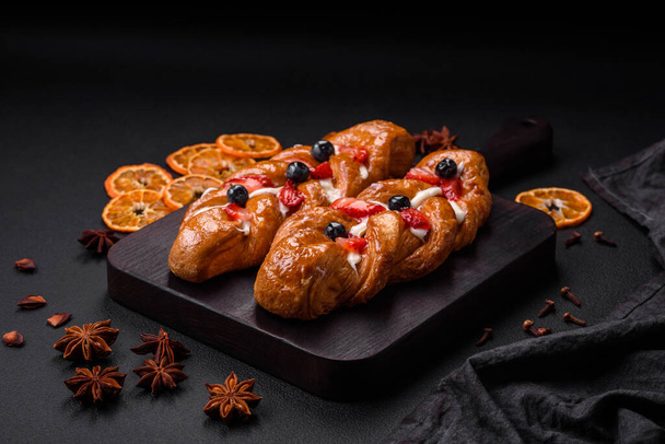 Delicious fresh crispy pastry in the form of a pigtail with strawberries and blueberries on a dark concrete background - 写真・画像